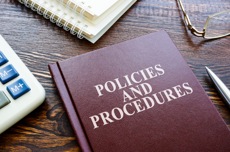 A book titled Policies and Procedures sits on a desk surrounded by a calculator, notepads, pen, eyeglasses, and other administration essentials.