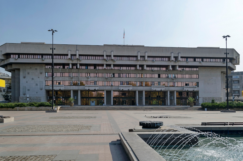 A large, rectangular administration or office building with multiple floors, flat roof, and rows of windows, situated behind an open plaza with a fountain in front.