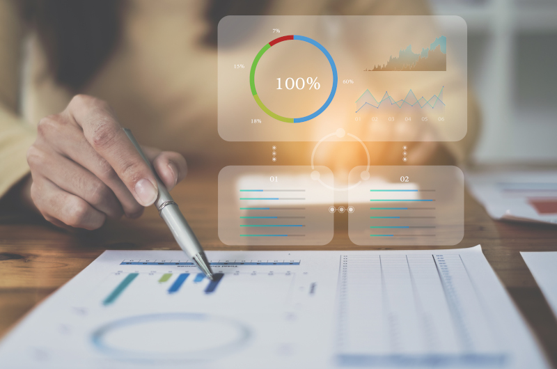 A person points at printed charts on a desk with a pen, showcasing effective administration as digital graphs and data visualizations are projected above the paper.