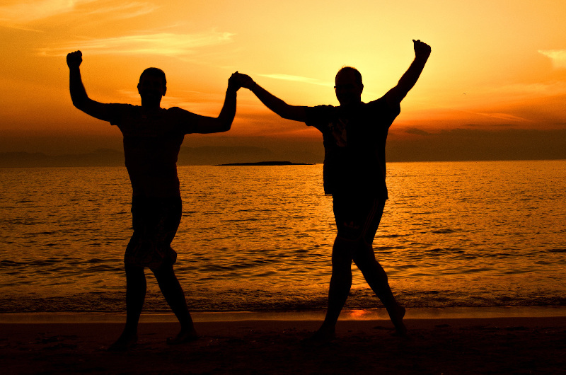 Two people hold hands and raise their arms while walking on a beach at sunset, silhouetted against the orange sky and calm sea—a perfect setting for a commercial celebrating togetherness.