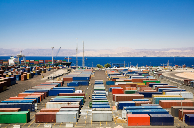 An industrial commercial port with colorful shipping containers arranged in rows near the sea under a clear blue sky. Cranes and infrastructure supporting active commerce are visible in the background.