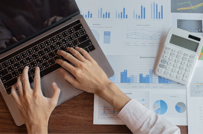 A person uses a laptop surrounded by printed charts, graphs, and a calculator, suggesting financial data analysis.