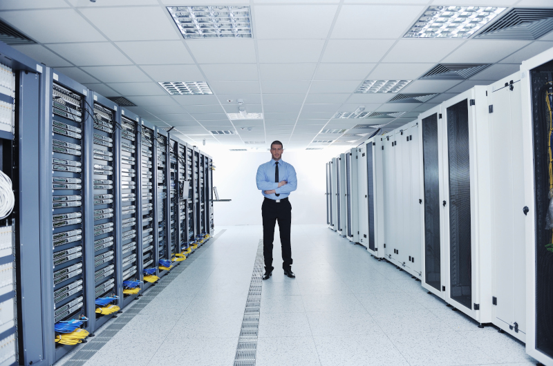 A man in business attire stands in the middle of a modern server room lined with server racks, overseeing critical information and network equipment.
