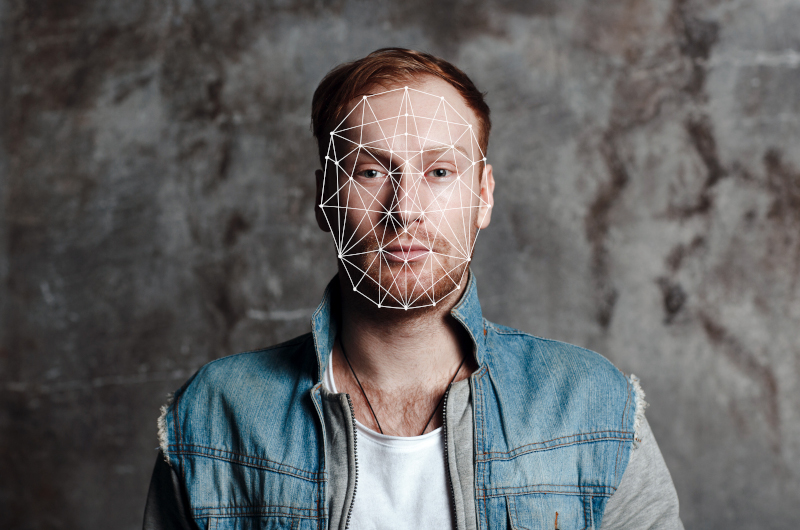 A person with a beard and red hair stands against a textured wall, with a geometric facial recognition grid overlayed on their face, highlighting how information can be mapped and analyzed through advanced technology.