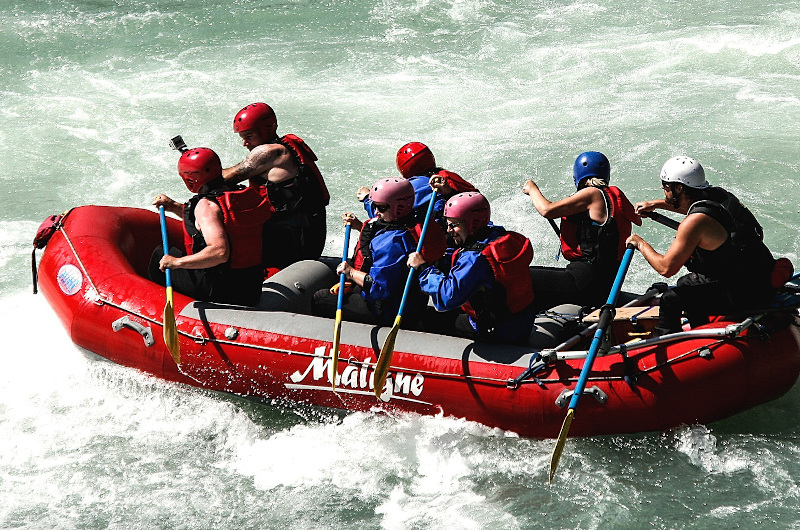 Six people from an organisation, wearing helmets and life jackets, paddle a red inflatable raft through whitewater rapids.
