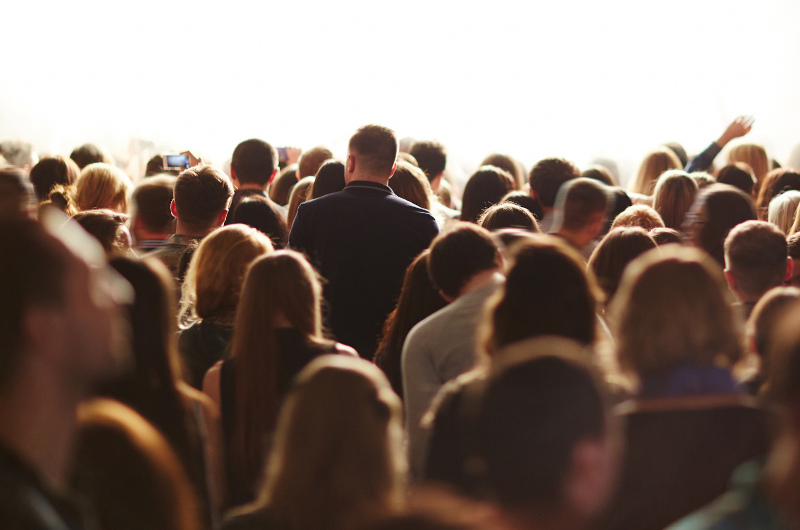 A large crowd of people stands facing a bright, white-lit area, with several people holding up phones to capture the scene.