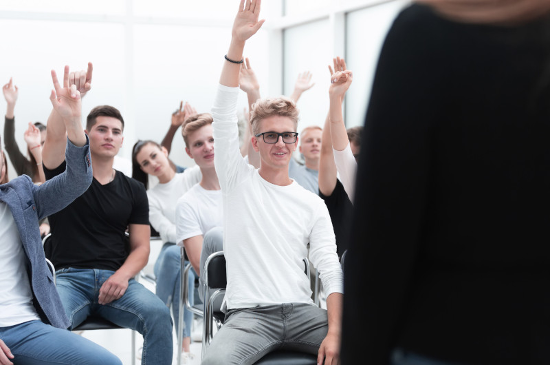 A group of students and people sit in a classroom, several with hands raised, actively participating in a discussion with a person standing at the front.