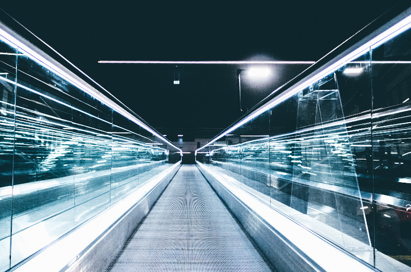 A moving walkway with glass sides stretches forward under bright lights in a modern, indoor setting, showcasing advanced technology amid strong lines and striking reflections.