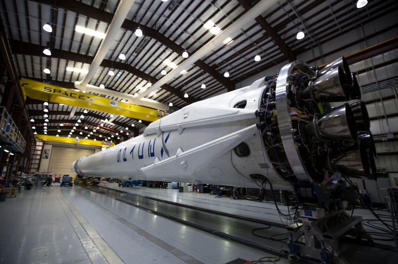 A SpaceX rocket booster is positioned horizontally inside a large industrial building, showcasing its engines, part of the company logo, and SpaceX's cutting-edge technology.
