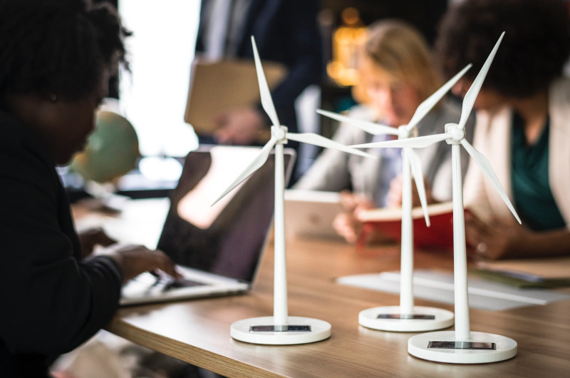 Three small wind turbine models sit on a table in a meeting room, showcasing innovative technology as people work and discuss in the background.