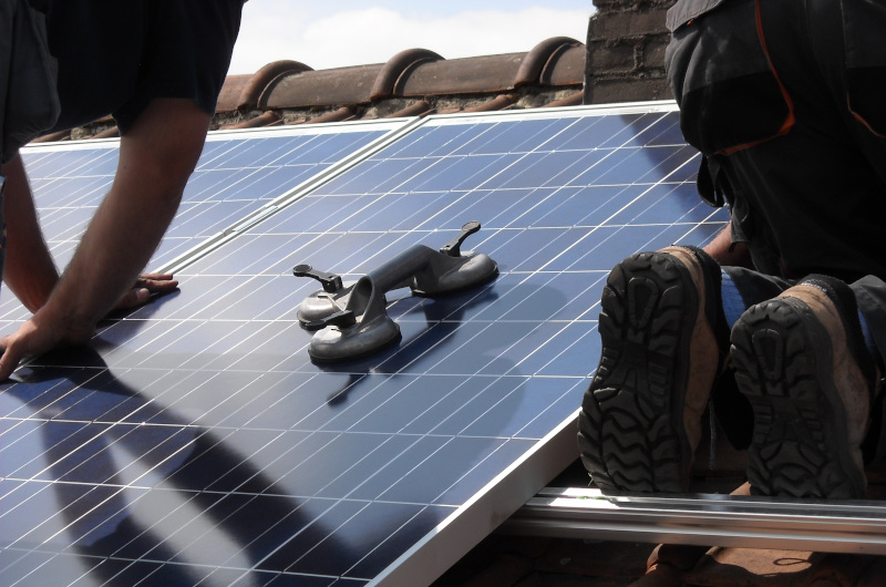 Two workers install solar panels on a rooftop, showcasing modern technology in action. A suction lifter tool rests on one of the panels, while only the workers' arms and legs are visible.